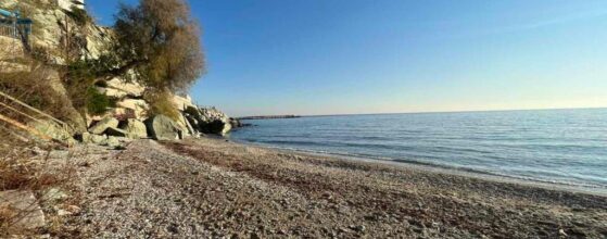 Ammoudeli Apartments - A rocky beach with gentle waves on a clear day. A tree and boulders are visible on the left side, with a bright, cloudless sky above. The coastline curves into the distance.