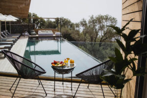 Five Olive Dream Trip - A serene wooden deck with two modern black chairs and a small round table holding fruit and juices, overlooks an infinity pool. Beyond the pool, lush trees and a misty horizon create a tranquil backdrop.