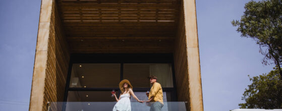 Five Olive Dream Trip - A couple stands on the glass balcony of a modern wooden cabin. The woman in a white dress and sunhat holds a drink, and the man in a yellow shirt and cap holds her hand. A clear blue sky and surrounding trees complete the scene.