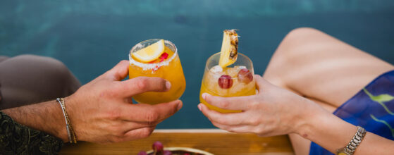Five Olive Dream Trip - Two people clink glasses of fruity cocktails topped with lemon and pineapple slices by a pool. A plate with grapes, strawberries, apples, and pineapple is on the wooden deck. The water in the background adds a refreshing ambiance.