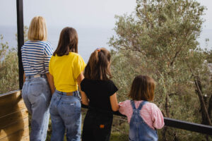 Five Olive Dream Trip - Four people stand on a balcony with their backs to the camera, overlooking a scenic view of trees and a distant body of water. They are dressed casually, and the atmosphere is calm and relaxed.