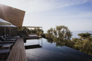 Five Olive Dream Trip - A serene infinity pool overlooking the ocean, surrounded by trees and a wooden deck with lounge chairs. The pool reflects the clear sky and nearby modern structure, creating a tranquil and luxurious setting.