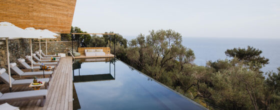 Five Olive Dream Trip - A modern outdoor infinity pool overlooks a scenic ocean view. The deck features several lounge chairs and umbrellas, with a small wooden pergola in the background. Lush greenery surrounds the space under a clear sky.