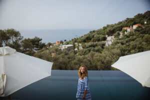 Five Olive Dream Trip - A person in a blue and white patterned dress stands by a modern infinity pool overlooking a scenic view of the ocean and lush green hills. Two large white umbrellas frame the foreground.