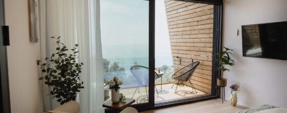 Five Olive Dream Trip - The Sunset Suite, a modern hotel room with floor-to-ceiling windows, offers stunning views of the sea. Inside, you'll find a cozy chair, lush plants, and a TV for your comfort. The balcony is equipped with two chairs and a small table, providing an inviting spot to enjoy the ocean view and admire the wood-paneled exterior.