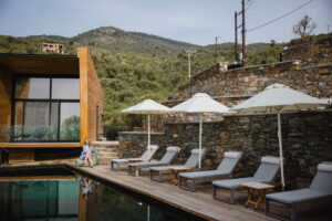 Five Olive Dream Trip - A serene outdoor poolside setting with lounge chairs and white umbrellas on a wooden deck. A person sits by the pool, surrounded by lush green hills and a modern stone building with large windows.