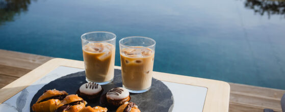 Five Olive Dream Trip - Two glasses of iced coffee sit on a wooden table beside an infinity pool with a scenic view. A slate platter holds several pastries, including chocolate-drizzled croissants and macarons.