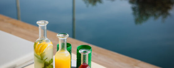 Five Olive Dream Trip - Bottles of yellow and red drinks with lemons and mint sit on a table next to green glasses overlooking a body of water.
