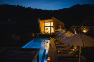 Five Olive Dream Trip - A modern cabin at dusk with large glass windows is illuminated from within. A long, narrow pool stretches alongside several lounge chairs with white umbrellas. The surrounding area is densely wooded with hills silhouetted against the evening sky.
