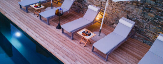 Five Olive Dream Trip - A serene poolside scene featuring four lounge chairs with cushions and small tables, under an umbrella. The pool is calm, and soft lighting enhances the stone wall and wooden deck, creating a tranquil atmosphere.