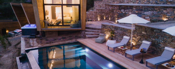 Five Olive Dream Trip - A modern hillside villa with a large illuminated window overlooks an outdoor pool. There's a wooden deck with loungers, tables, and umbrellas beside the pool, set against a backdrop of stone walls and green hills. The scene is lit by warm lights.