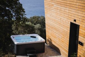 Five Olive Dream Trip - The Sunset Suite features a cozy jacuzzi on a stone patio next to a wooden building. It's nestled among trees and offers views of the water in the background, creating a tranquil and soothing environment.