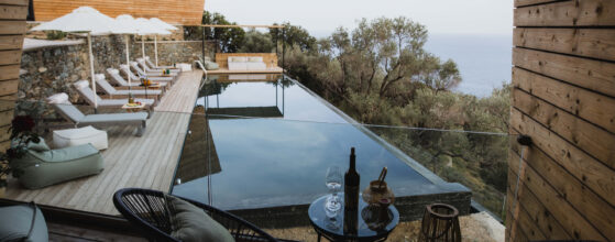 Five Olive Dream Trip - The Freedom Suite features a tranquil patio that overlooks an infinity pool with lounge chairs shaded by umbrellas. Beyond the pool lies a panoramic view of lush greenery and the sea. In the foreground, there is a cozy seating area equipped with two chairs and a small table adorned with wine glasses.