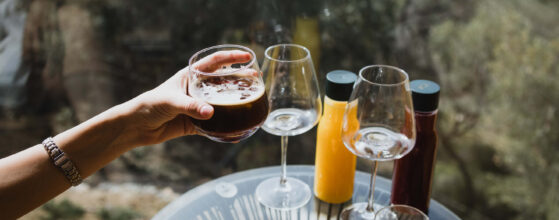 Five Olive Dream Trip - In the Honeymoon Suite, a hand holds a glass of iced coffee in focus, while the background features a table set for drinks with two empty wine glasses, tall yellow and red bottles, and a plate with biscuits and snacks. A scenic view of trees and water can be seen outside.