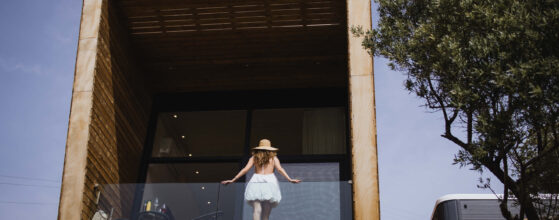 Five Olive Dream Trip - A woman in a white dress and straw hat stands on the balcony of the Honeymoon Suite, gazing at the view. The modern wooden building boasts large glass panels and is nestled amidst rocky terrain with a tree to the right.