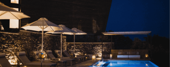 Five Olive Dream Trip - A nighttime pool scene with lounge chairs and umbrellas lined along the deck exudes the charm of Plomari. Soft, ambient lighting illuminates the stone wall and surrounding area, creating a cozy and inviting atmosphere. The dark sky enhances the tranquil mood.