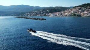NRN Homeland Greece - Aerial view of a small motorboat leaving a foamy wake as it speeds across a deep blue sea near Plomari. In the background, this coastal town with clustered buildings sits at the base of green hills, adjacent to a long stone pier.