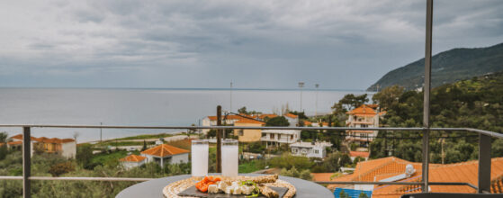A round table on a balcony holds two glasses and a plate of appetizers, overlooking a coastal village with orange-roofed houses, greenery, and the sea under a cloudy sky.