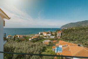 A scenic coastal village with red-roofed houses surrounded by green hills overlooks a calm blue sea under a clear sky, with mountains visible in the distance.