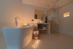 A modern, minimalist bathroom with a white freestanding bathtub, lit candles on the floor and counter, rolled towels in baskets, potted plants, and a walk-in shower with a glass partition. The lighting is warm and relaxing.