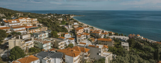 A coastal village with white buildings and terracotta roofs sits next to lush greenery, overlooking a calm, blue sea under a partly cloudy sky.