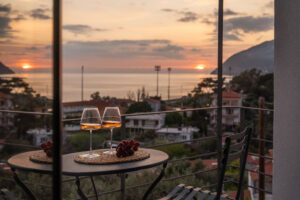 A small table with two glasses of wine and bunches of grapes sits on a balcony overlooking rooftops and the sea at sunset, with mountains and a partly cloudy sky in the background.