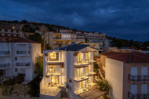 A modern, three-story building with large illuminated windows stands among other houses on a hillside at dusk, under a cloudy evening sky.