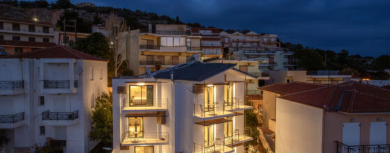 A modern, three-story building with large illuminated windows stands among other houses on a hillside at dusk, under a cloudy evening sky.