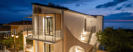 A modern white two-story house with outdoor lighting, large windows, and curved staircase, set against a twilight sky near the sea. Trees and a streetlamp are visible in the foreground.
