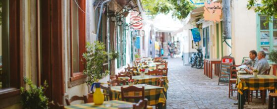 A narrow cobblestone street lined with tables and chairs covered in yellow plaid tablecloths, leading to a cozy outdoor café shaded by leafy green trees, with two people seated in the distance.