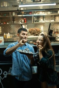 A man and woman stand in front of a food truck, smiling and sharing skewered food. The man holds a tray while both enjoy their food, with the truck’s kitchen and rotating meat visible in the background.