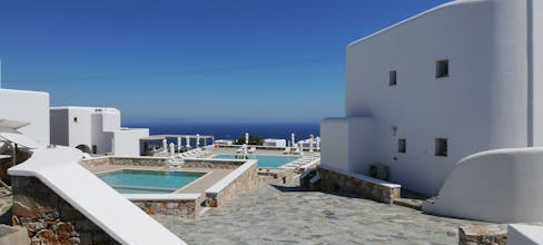 A sunny courtyard with white-washed buildings, a stone-tiled ground, a swimming pool, and a view of the deep blue sea under a clear sky, framed by an arched entryway.
