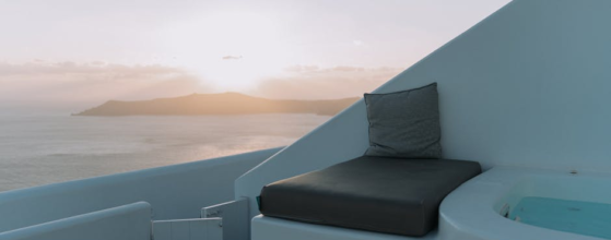 Een minimalistische lounge in de open lucht, met een stoel met kussens en een klein zwembad, kijkt uit over een rustige zee bij zonsondergang, met in de verte bergen en een pastelkleurige lucht op de achtergrond.