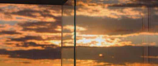 Golden sunset over the ocean, viewed through clear glass railings, with dramatic clouds scattered across the sky and warm orange and yellow tones reflecting on the water.
