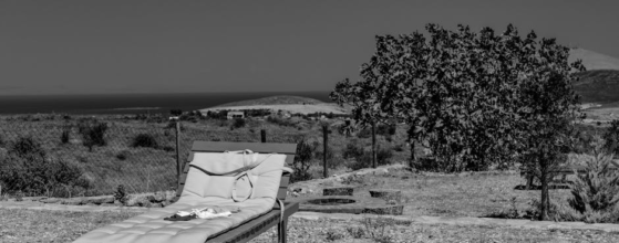 A black and white photo of a lounge chair with a towel and hat on it, placed on a pebbled ground outdoors. In the background, there's a tree, a fence, and a distant view of hills and water under a clear sky.
