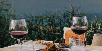 A wooden table set with wine glasses, water glasses, and plates of food overlooks a scenic body of water and distant hills, creating a peaceful outdoor dining scene.