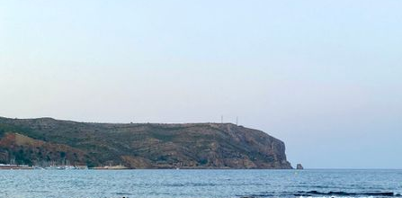 Rocky shoreline with scattered pebbles, calm blue sea, and a distant cliff under a clear sky. The coast extends into the water, creating a serene coastal landscape.