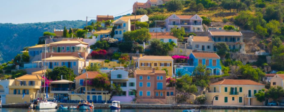 Colorful houses with orange roofs on a hillside overlook a calm bay with sailboats and clear water, under a bright blue sky.