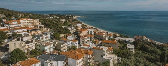 Aerial view of white buildings with terracotta roofs clustered on a hillside overlooking a calm sea and beach.